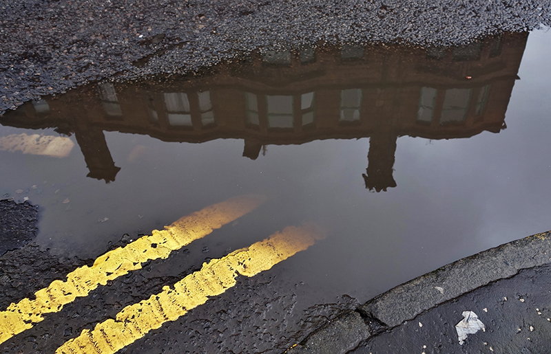 A tenement in a Glasgow street reflected in a puddle with double yellow lines on the road